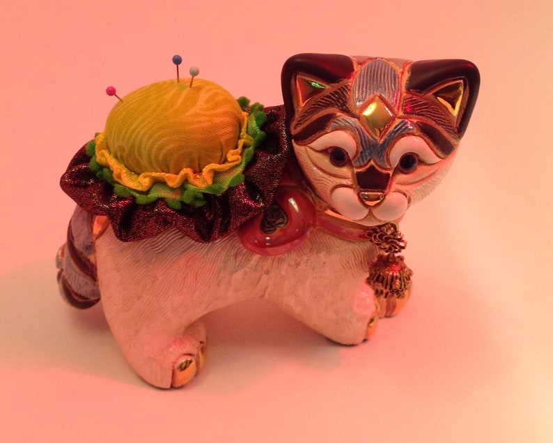 May include: A white ceramic cat figurine with black and blue markings on its face. The cat is wearing a red and green ribbon and has a yellow pincushion on its back. The pincushion has several colorful pins sticking out of it.