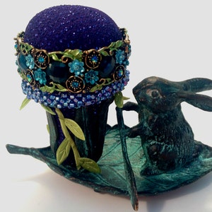 Puede incluir: Un cojín de alfileres morado con un ribete de cuentas azules y verdes se asienta sobre una base de metal verde en forma de hoja con una figurita de conejo negro en la parte superior.