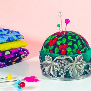 May include: A round pincushion with a green and black fabric cover with red berries and green leaves. The pincushion is sitting on a silver base with a grapevine design. There are three pins stuck in the pincushion.