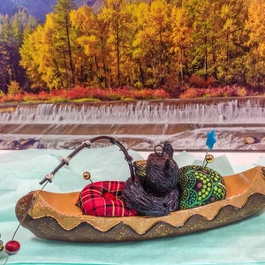 Puede incluir: Un cojín de alfileres de color marrón con forma de canoa con una figurita de oso negro, un cojín de alfileres de cuadros rojos y negros y un cojín de alfileres verde con una estrella azul en la parte superior. El cojín de alfileres está sobre una superficie azul con una imagen de una cascada y follaje de otoño en el fondo.