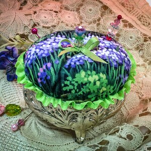 May include: A green and purple floral pincushion with a green ribbon and a silver stand with three legs. The pincushion is decorated with beads and pins.