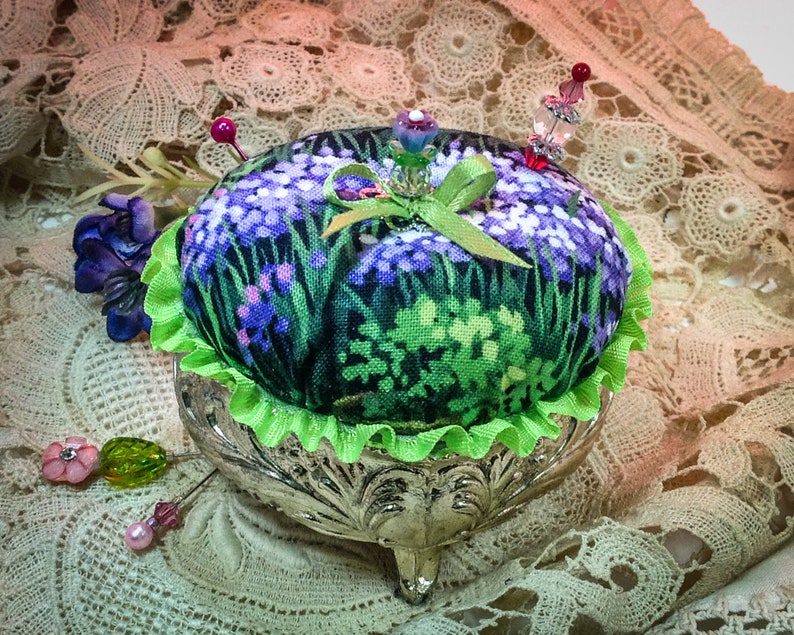 May include: A green and purple floral pincushion with a green ribbon and a silver stand with three legs. The pincushion is decorated with beads and pins.
