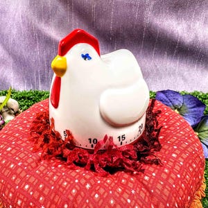 May include: A white ceramic chicken-shaped kitchen timer with a bright red comb, a yellow beak, and a blue eye detail. The base of the timer features black printed numbers ranging from 5 to 20, indicating time increments in minutes for cooking tasks.