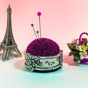 May include: A silver pincushion with a purple velvet top and a grapevine design. The pincushion has the word "Vin" inscribed on it. There are three pins sticking out of the top, one pink, one green, and one dark green. A miniature Eiffel Tower and a small purple basket with pink and yellow flowers are in the background.
