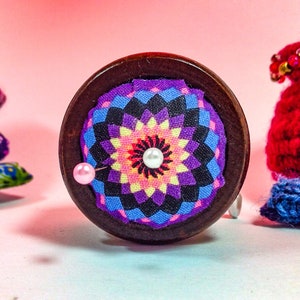 May include: A wooden pincushion with a colorful floral fabric top. The pincushion has a pink pin with a pearl head.