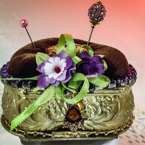 Op de afbeelding: Een gouden naaldenkussen met een bruine fluwelen bovenkant. Het naaldenkussen is versierd met paarse en witte bloemen, groen lint en strass. Er zitten twee naalden met roze en paarse glazen kralen bovenop.