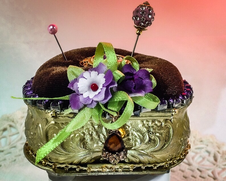Op de afbeelding: Een gouden naaldenkussen met een bruine fluwelen bovenkant. Het naaldenkussen is versierd met paarse en witte bloemen, groen lint en strass. Er zitten twee naalden met roze en paarse glazen kralen bovenop.