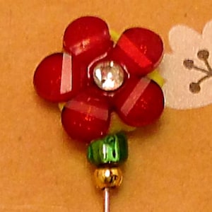 May include: A flower-shaped pin with red petals, white stripes, and a clear gem center. The pin has a green and gold stem. The background is a light orange color.