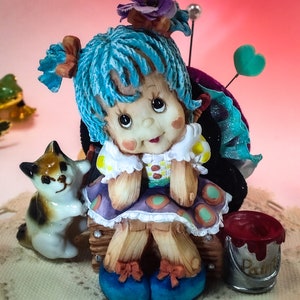 May include: A blue haired doll with a cat and a paint can. The doll is sitting on a wooden stool and has a pincushion on her back. The doll is wearing a purple and white dress with polka dots.