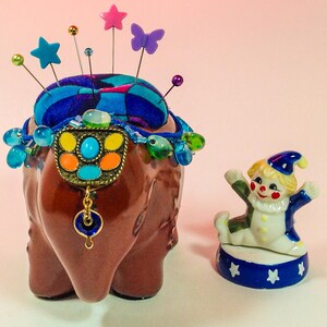 May include: A brown ceramic elephant pincushion with a blue and green patterned fabric top. The pincushion is decorated with colourful beads and a metal charm. A small porcelain clown figurine is standing on a blue and white base in front of the pincushion.