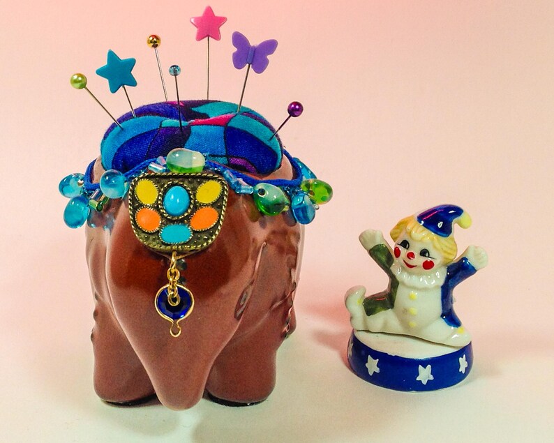 May include: A brown ceramic elephant pincushion with a blue and green patterned fabric top. The pincushion is decorated with colourful beads and a metal charm. A small porcelain clown figurine is standing on a blue and white base in front of the pincushion.