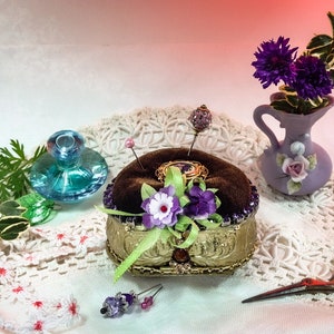 Op de afbeelding: Een vintage-stijl naaldenkussen met een fluwelen bovenkant en gouden versiering. Het naaldenkussen is versierd met paarse en witte bloemen en een groen lint. Het staat op een witte kantonderlegger met een klein blauw glazen flesje en een vaas met paarse bloemen.