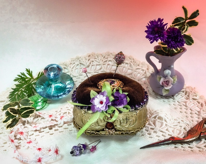 Op de afbeelding: Een vintage-stijl naaldenkussen met een fluwelen bovenkant en gouden versiering. Het naaldenkussen is versierd met paarse en witte bloemen en een groen lint. Het staat op een witte kantonderlegger met een klein blauw glazen flesje en een vaas met paarse bloemen.
