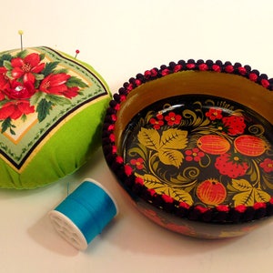 May include: A green pincushion with red floral print and a black and gold bowl with a red and yellow floral design. The bowl has a black and red trim.