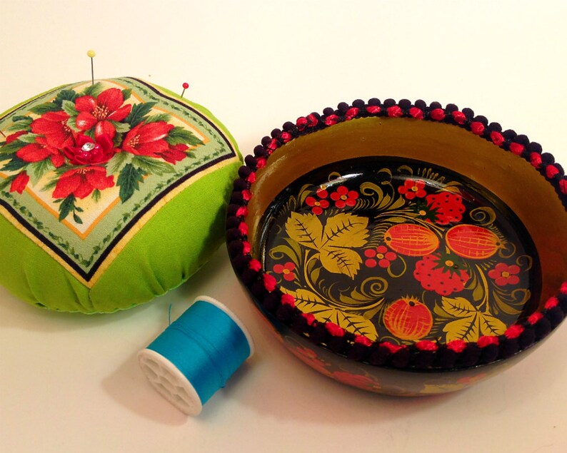 May include: A green pincushion with red floral print and a black and gold bowl with a red and yellow floral design. The bowl has a black and red trim.