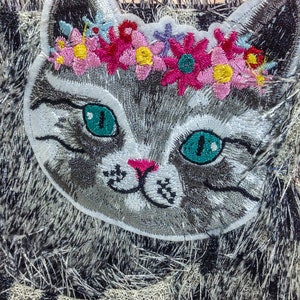 May include: A gray embroidered cat with green eyes and a pink nose wearing a flower crown with pink, yellow, and blue flowers. The cat is on a black and white fuzzy background.