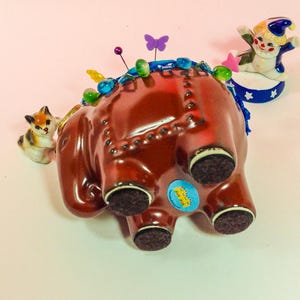 May include: A brown ceramic elephant pincushion with blue and green trim, a small cat figurine, and a clown figurine on a blue and white platform. The elephant has a blue ribbon around its trunk and a variety of colourful pins stuck in its back.