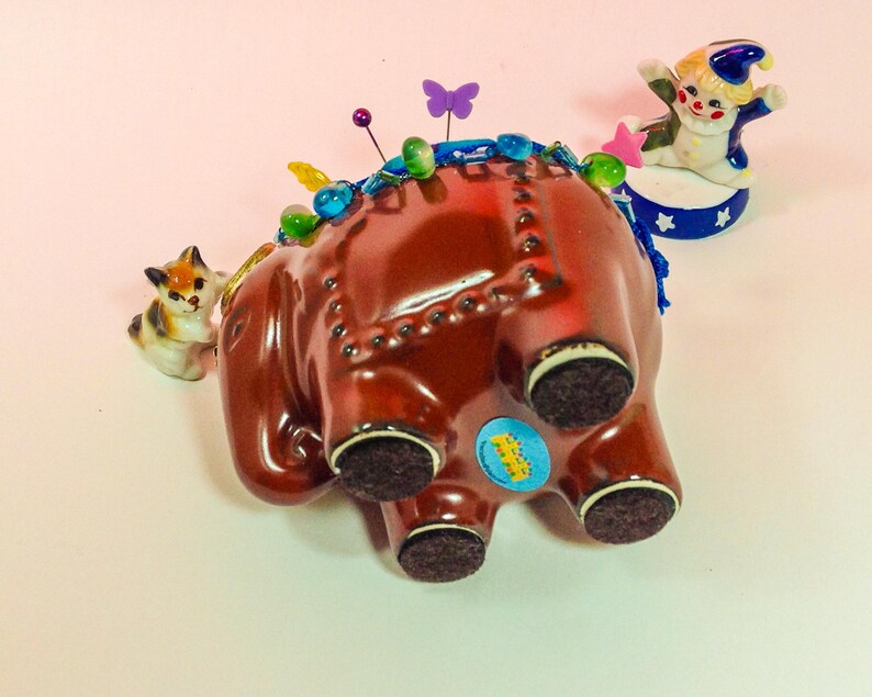 May include: A brown ceramic elephant pincushion with blue and green trim, a small cat figurine, and a clown figurine on a blue and white platform. The elephant has a blue ribbon around its trunk and a variety of colourful pins stuck in its back.