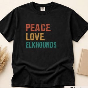 Può includere: Una t-shirt nera a maniche corte con il testo PEACE. LOVE. ELKHOUNDS. stampato in un carattere dall'aspetto vissuto. La parola PEACE è arancione, LOVE è gialla ed ELKHOUNDS è verde acqua. La maglia ha un girocollo e un'etichetta Comfort Colors.