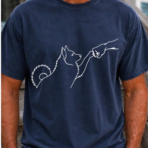 以下が含まれることがあります： 胸元に白いラインアートのグラフィックが施されたネイビーブルーの半袖Tシャツ。デザインは、ふさふさした毛並みと巻いた尻尾を持つ犬のシルエットが、人間の手と拳を合わせるポーズをとっている様子をシンプルな線で描いています。