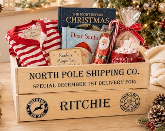 May include: A wooden crate labeled North Pole Shipping Co. for Ritchie, containing red and white striped pajamas, The Night Before Christmas book, Dear Santa book, a Santa's Magic Key sign, a candy cane, and a mug filled with marshmallows.
