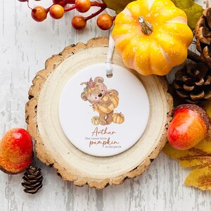 May include: A round ceramic ornament featuring a watercolour illustration of a brown bear wearing a pumpkin costume. The text "Arthur The cutest little pumpkin in the patch" is written below the bear.