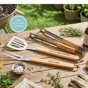 May include: A collection of barbecue tools with wooden handles and stainless steel heads, including a spatula, fork, and tongs. The handles are engraved with phrases like "Happy Father's Day" and "King of the BBQ!" on a wooden table.