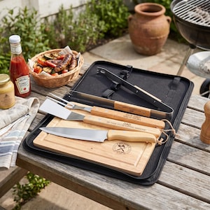 May include: A barbecue set with wooden-handled tools, including a knife, spatula, and fork, presented in a black case. A wooden chopping board is included. Ketchup, mustard, and a basket of grilled food are also on the table.