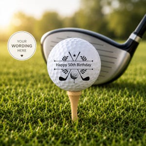May include: A white golf ball resting on a wooden tee, featuring a black graphic of crossed golf clubs, a small golf ball, and laurel branches with the text "Happy 50th Birthday" printed in the centre. A golf club head is visible behind the ball.