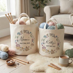 May include: Two off-white canvas storage bags with two handles each, featuring a printed wreath of knitting supplies and the text Nana's KNITTING BAG and Kathy's KNITTING BAG. The bags are filled with colourful yarn balls and wooden knitting needles.