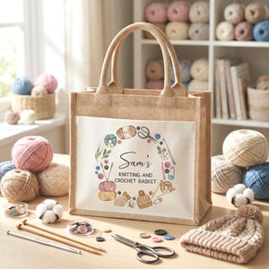 May include: A beige jute tote bag with a white front pocket featuring a watercolour wreath design with knitting and crochet tools. The text "Sam's Knitting and Crochet Basket" is in the centre. Knitting needles, yarn, and a knitted cap are on the table.