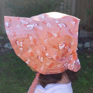 May include: A peach-colored bonnet with a wide brim, featuring a repeating pattern of white and orange floral designs. The bonnet is tied with a matching ribbon. The hat is made of a lightweight fabric, suitable for sun protection.