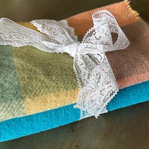 May include: A folded textile piece featuring a multi-coloured plaid pattern in shades of sage green, yellow, and peach, layered atop a solid bright turquoise textile. The two pieces are tied together with a white floral lace ribbon bow.