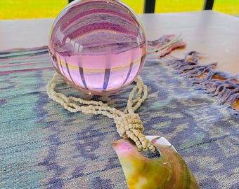 Puede incluir: Una esfera de vidrio transparente refleja un interior rosa y morado. Un collar de cuentas color crema con un colgante de concha descansa sobre un textil estampado azul y rosa. El colgante es iridiscente con tonos verdes, rosas y marrones.