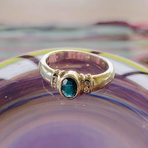 May include: Silver ring with an oval, teal gemstone as the focal point. The ring has a plain band with decorative elements either side of the central stone, each with small, clear stones. The ring is set against a blurred, colourful background.