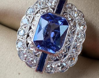 May include: A vintage-style ring featuring a large, faceted, blue sapphire surrounded by a halo of smaller diamonds. The ring is set in silver and has a delicate, intricate design.