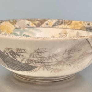 May include: Off-white ceramic bowl with a hand-painted landscape design. The bowl features a rim with geometric patterns in yellow, brown, and gray. The interior depicts a mountain and bamboo scene in shades of gray and black.