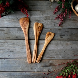 May include: Three wooden kitchen spatulas of varying sizes, made from olive wood, with natural wood grain patterns. The spatulas are arranged on a rustic wooden surface.