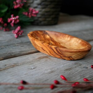May include: A small, oval-shaped wooden bowl made from olive wood. The bowl has a natural wood grain pattern and is light brown in color.