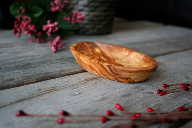 May include: A small, oval-shaped wooden bowl made from olive wood. The bowl has a natural wood grain pattern and is light brown in color.
