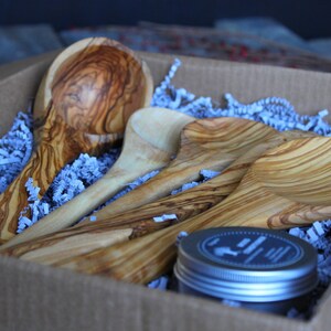 Puede incluir: Un juego de cuatro cucharas de madera y un peque&ntilde;o frasco de acondicionador para madera en una caja de cart&oacute;n marr&oacute;n con papel de seda azul. Las cucharas est&aacute;n hechas de madera de olivo y tienen un acabado natural.