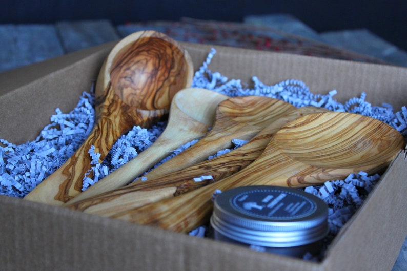 Puede incluir: Un juego de cuatro cucharas de madera y un peque&ntilde;o frasco de acondicionador para madera en una caja de cart&oacute;n marr&oacute;n con papel de seda azul. Las cucharas est&aacute;n hechas de madera de olivo y tienen un acabado natural.