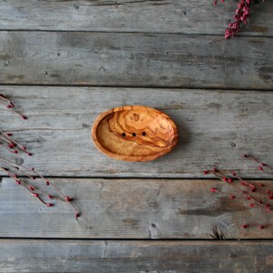 May include: A wooden soap dish with a natural wood grain pattern and three small holes for drainage. The dish is oval shaped and is sitting on a wooden surface.