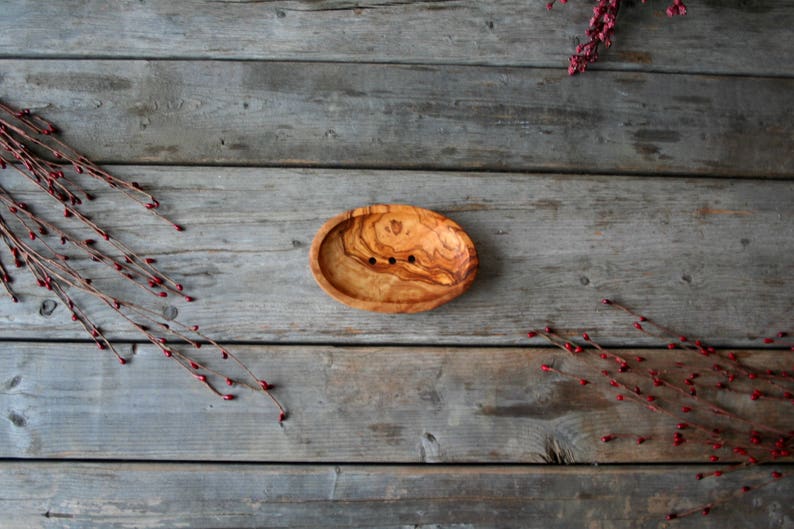 May include: A wooden soap dish with a natural wood grain pattern and three small holes for drainage. The dish is oval shaped and is sitting on a wooden surface.