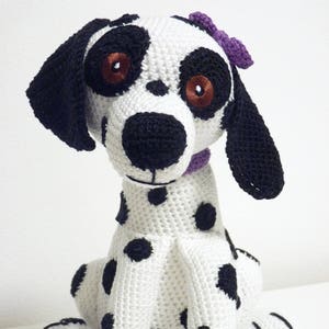 Puede incluir: Un juguete de peluche de dálmata tejido a crochet con manchas negras, ojos marrones y un lazo morado. El perro está sentado con las patas metidas debajo.