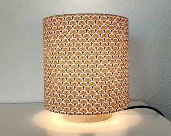 May include: A table lamp with a white and gold patterned lampshade. The lampshade has a geometric design of overlapping fans. The lamp has a wooden base.