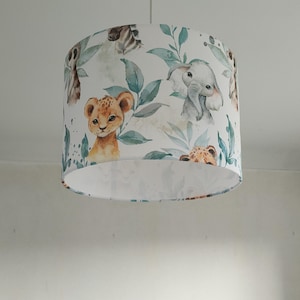 Puede incluir: Pantalla de lámpara cilíndrica blanca con un diseño de acuarela de animales de la jungla, incluyendo un cachorro de león, un elefante y una cebra, entre follaje verde. Ideal para una habitación infantil, cuelga de un cable blanco.