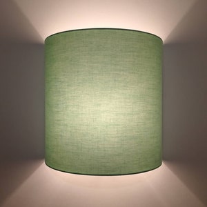 Op de afbeelding: Een cilindrische wandlamp met een lichtgroene linnen kap. De lamp is aan een witte muur bevestigd en werpt een zachte gloed boven en onder. De kap heeft een donkergroene rand.