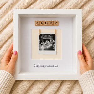 May include: A white square shadow box frame containing a black and white ultrasound image clipped to a cream-coloured backing. Above the image are wooden letter tiles spelling DADDY. Below the image is the text I can't wait to meet you!