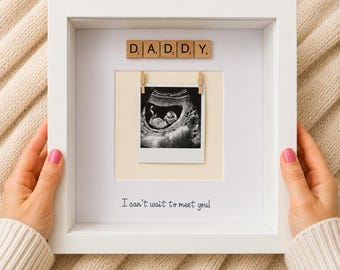 May include: A white square shadow box frame containing a black and white ultrasound image clipped to a cream-colored backing. Above the image are wooden letter tiles spelling DADDY. Below the image is the text I can't wait to meet you!